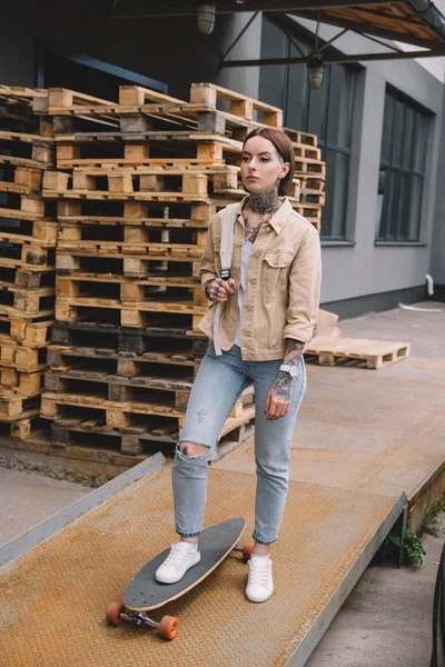 Elegante tatuado mulher de pé com skate perto de paletes de madeira — Fotografia de Stock