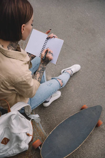 Vista de ángulo alto de mujer tatuada con escritura de monopatín en libro de texto vacío - foto de stock