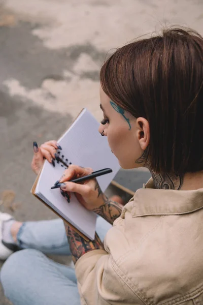 Vista de ángulo alto de la mujer joven tatuada escribiendo en cuaderno en blanco - foto de stock