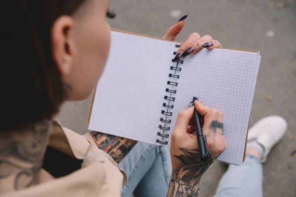 Foco seletivo da jovem tatuada escrevendo em livro didático vazio — Fotografia de Stock