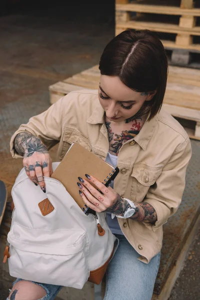 Visão de alto ângulo de mulher tatuada elegante colocando livro didático na mochila — Fotografia de Stock