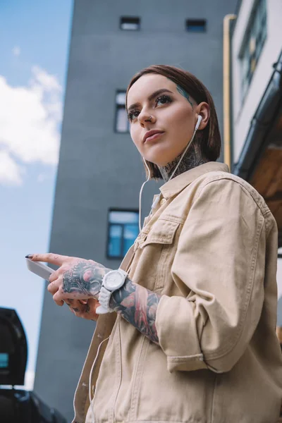 Vista a basso angolo della donna tatuata in auricolare ascoltare musica con smartphone — Foto stock