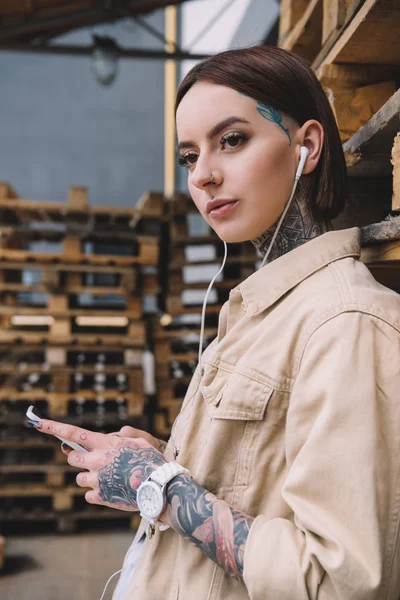 Joven mujer tatuada en auriculares escuchando música con smartphone - foto de stock
