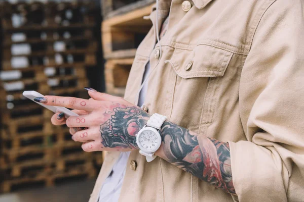 Imagen recortada de mujer tatuada con reloj de pulsera usando teléfono inteligente - foto de stock