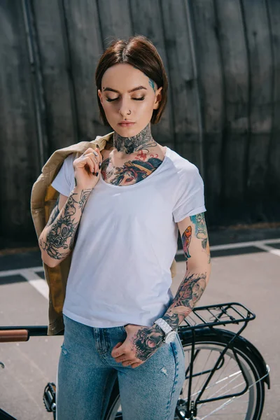 Joven mujer tatuada con los ojos cerrados sosteniendo chaqueta sobre el hombro cerca de la bicicleta - foto de stock