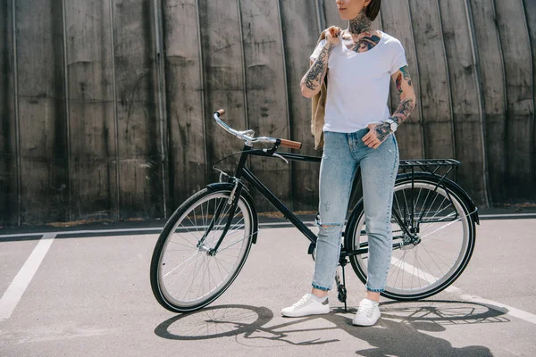 Imagem cortada de mulher tatuada elegante segurando jaqueta sobre ombro perto de bicicleta na rua — Fotografia de Stock