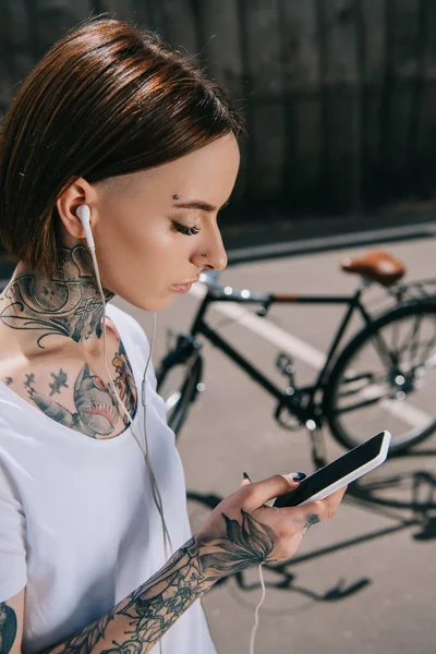 Vista laterale della donna tatuata in auricolare ascoltare musica con smartphone vicino alla bicicletta — Foto stock