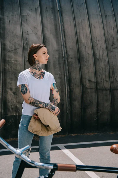 Vista lateral de mujer con estilo con tatuajes mirando lejos cerca de la bicicleta - foto de stock