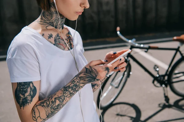 Cropped image of tattooed woman in earphones listening music with smartphone near bicycle — Stock Photo