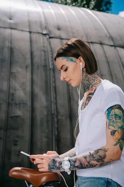 Mujer tatuada sonriente en auriculares escuchando música con teléfono inteligente mientras está sentada en bicicleta - foto de stock