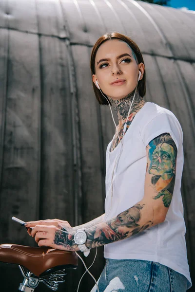 Low angle view of tattooed woman in earphones listening music with smartphone on bicycle — Stock Photo