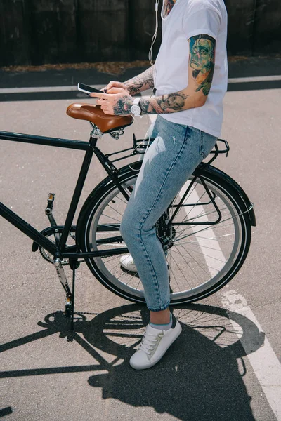 Cropped image of tattooed woman in earphones listening music with smartphone while sitting on bicycle — Stock Photo
