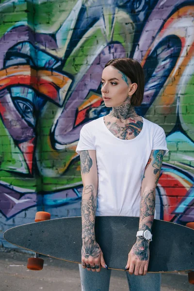 Jeune fille avec des tatouages tenant longboard contre le mur avec des graffitis colorés — Photo de stock