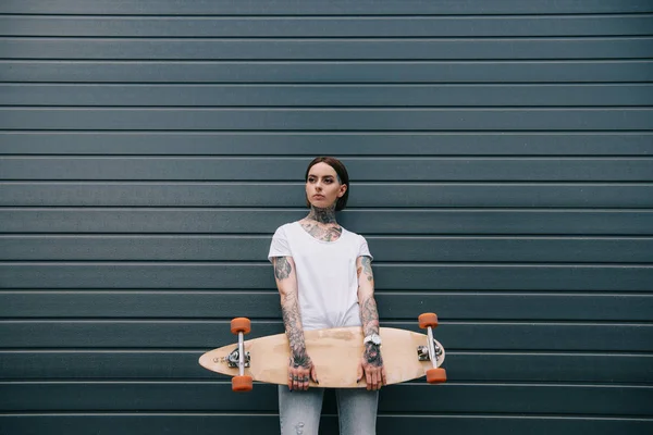 Attractive young woman with tattoos holding skateboard against black wall — Stock Photo