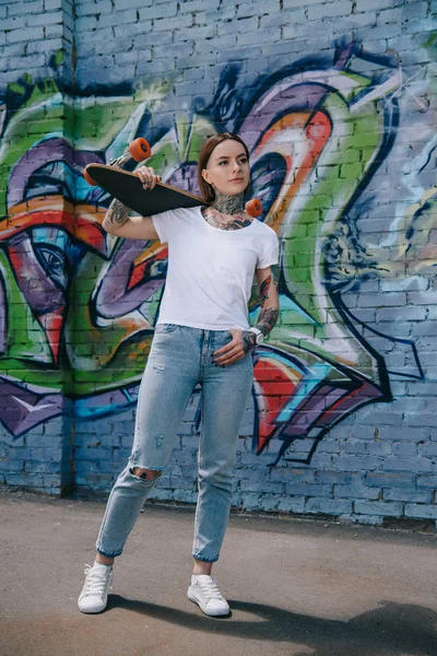 Jolie fille avec des tatouages tenant skateboard sur l'épaule près du mur avec des graffitis — Photo de stock