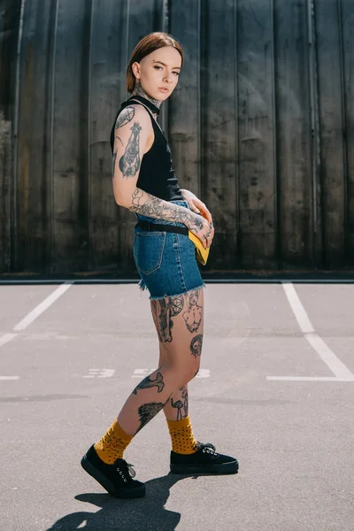 Atractiva joven mujer tatuada con bolsa de cintura mirando a la cámara - foto de stock