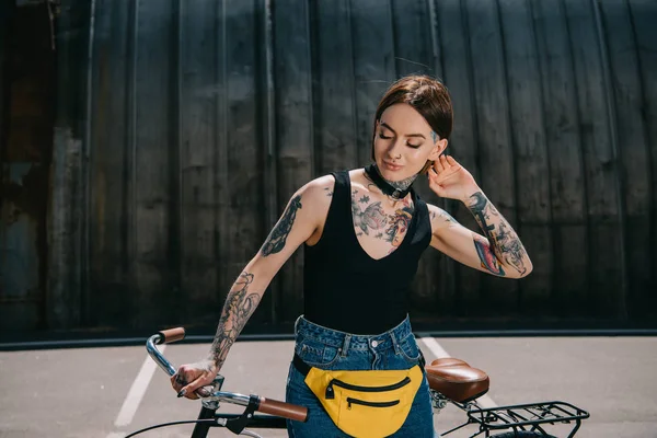 Улыбающаяся стильная татуированная девушка с закрытыми глазами стоящая с велосипедом на улице — стоковое фото