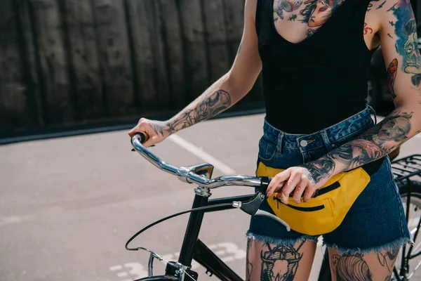 Обрезанный образ стильной татуированной девушки, стоящей с велосипедом на улице — стоковое фото