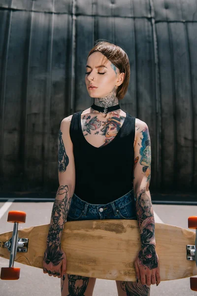 Atractiva chica tatuada con estilo con los ojos cerrados sosteniendo monopatín - foto de stock