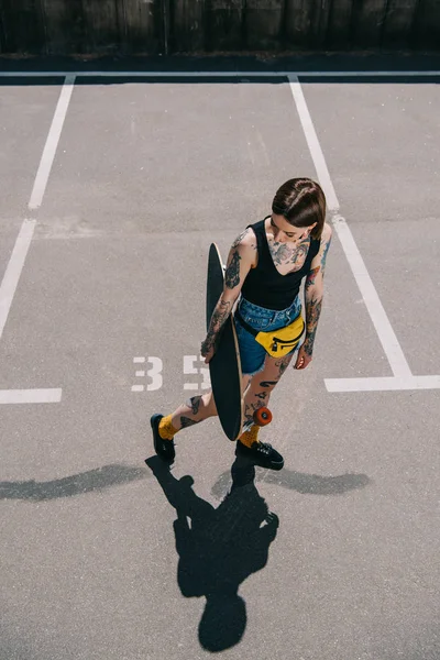 Visão de alto ângulo de elegante menina tatuada segurando skate no estacionamento — Fotografia de Stock