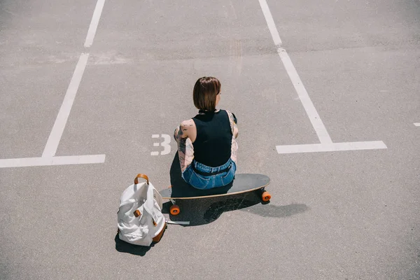 Visão traseira da menina tatuada elegante sentado no skate perto da mochila no estacionamento — Fotografia de Stock