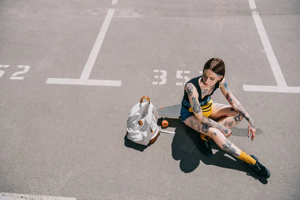 Vista ad alto angolo di elegante ragazza tatuata seduta sullo skateboard vicino allo zaino nel parcheggio — Foto stock