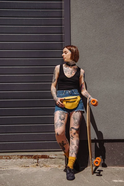 Élégante fille tatouée avec sac de taille tenant skateboard contre mur — Photo de stock