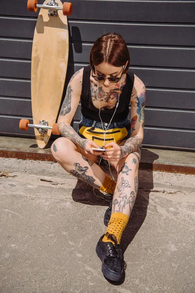 Young tattooed woman in earphones listening music with smartphone near skateboard at street — Stock Photo