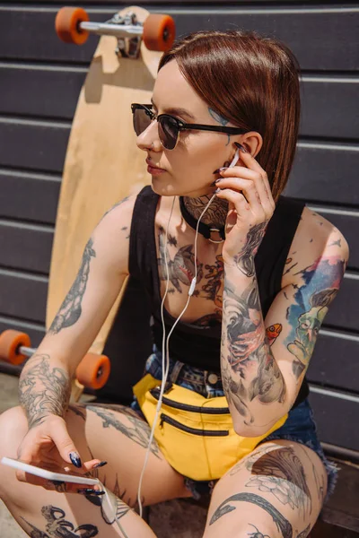 Tattooed woman in earphones listening music with smartphone near skateboard — Stock Photo