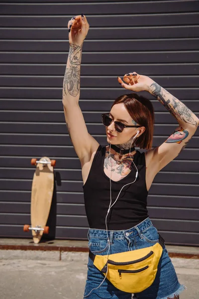 Stylish tattooed girl in earphones listening music while standing with raised arms — Stock Photo