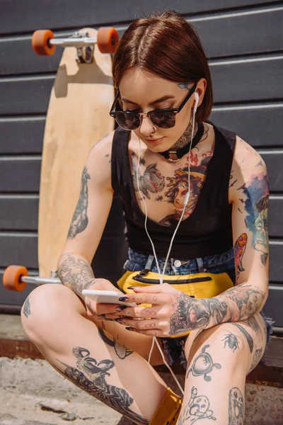 Молодая татуированная женщина в наушниках слушает музыку со смартфоном возле скейтборда — стоковое фото