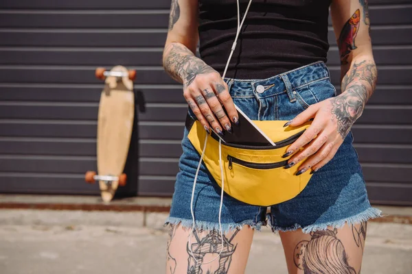 Image recadrée de fille tatouée mettant smartphone avec écran vide dans le sac de taille — Photo de stock