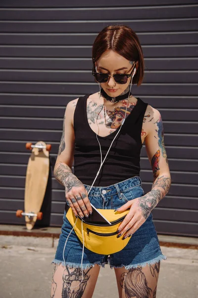 Stylish tattooed woman listening music in earphones and putting smartphone in waist bag — Stock Photo