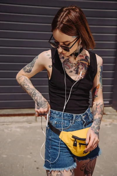 Élégante fille tatouée écouter de la musique dans les écouteurs et mettre smartphone dans la poche — Photo de stock