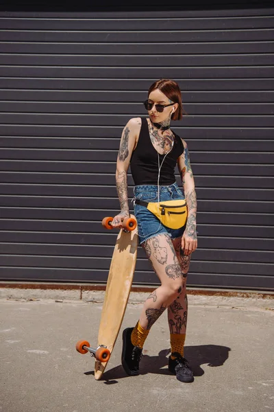 Femme tatouée élégante dans des lunettes de soleil écouter de la musique avec des écouteurs et debout près de planche à roulettes — Photo de stock