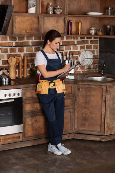 Selbstbewusste junge Reparaturfrau schreibt in Klemmbrett, während sie in der Küche steht — Stockfoto