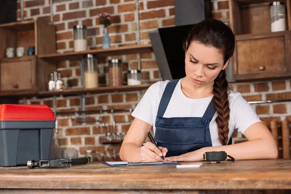 Confiado joven reparadora en la escritura general en portapapeles en la cocina — Stock Photo