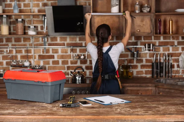 Various tools lying on kitchen table with blurred young repairwoman measuring cabinet on background — Stock Photo