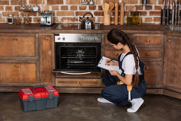 Молодая ремонтница осматривает кухонную печь и пишет в буфер обмена — стоковое фото