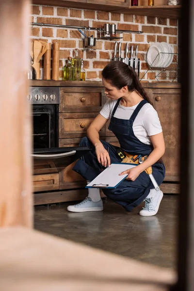 Serious young repairwoman with clipboard sitting near oven and looking at it — Stock Photo