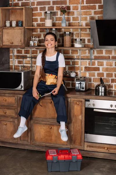 Feliz jovem reparadora em uniforme sentado na mesa da cozinha e olhando para a câmera — Fotografia de Stock