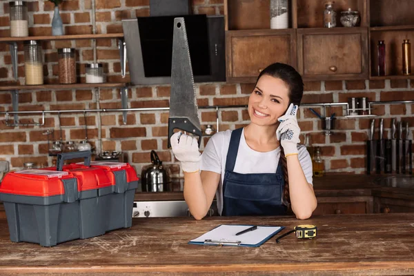 Felice giovane riparatrice con sega presa per telefono in cucina — Foto stock