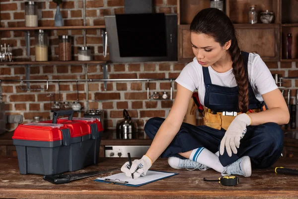 Belle jeune réparatrice assise sur la table de cuisine avec des outils et de l'écriture dans le presse-papiers — Photo de stock