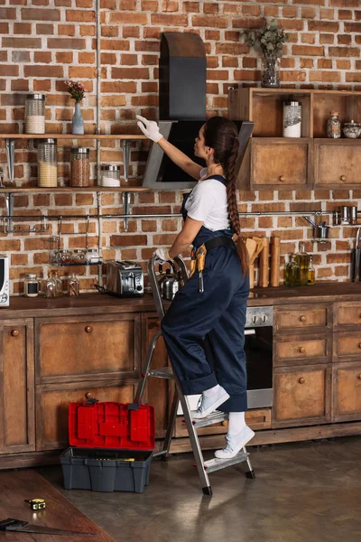 Beautiful young repairwoman standing on stepladder at kitchen — Stock Photo