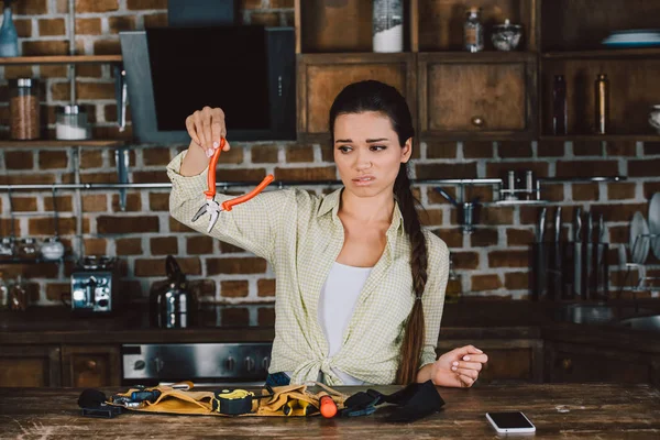 Confused young woman looking at pliers at kitchen — Stock Photo