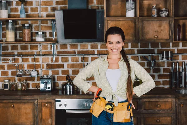 Glücklich junge Reparaturfrau mit Armen akimbo steht in der Küche und blickt in die Kamera — Stockfoto