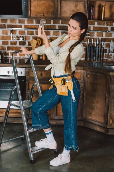 Hermosa joven reparadora de pie en la escalera y examinar la bombilla en la cocina - foto de stock