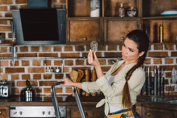 Confident young repairwoman standing on stepladder and examining light bulb at kitchen — Stock Photo