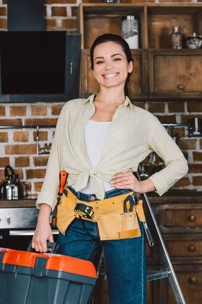 Felice giovane riparatrice con cintura degli attrezzi e scatola in piedi in cucina e guardando la fotocamera — Foto stock