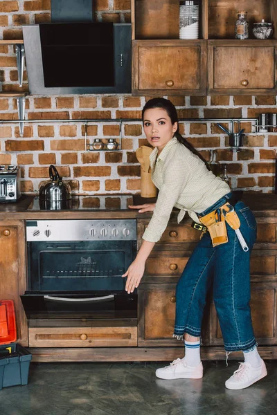 Geschockte junge Reparaturfrau öffnet kaputten Ofen mit Rauch im Inneren — Stockfoto